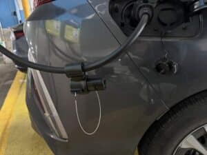 A close-up of a gray electric car being charged at a charging station, with the charging cable plugged into the vehicle's port.