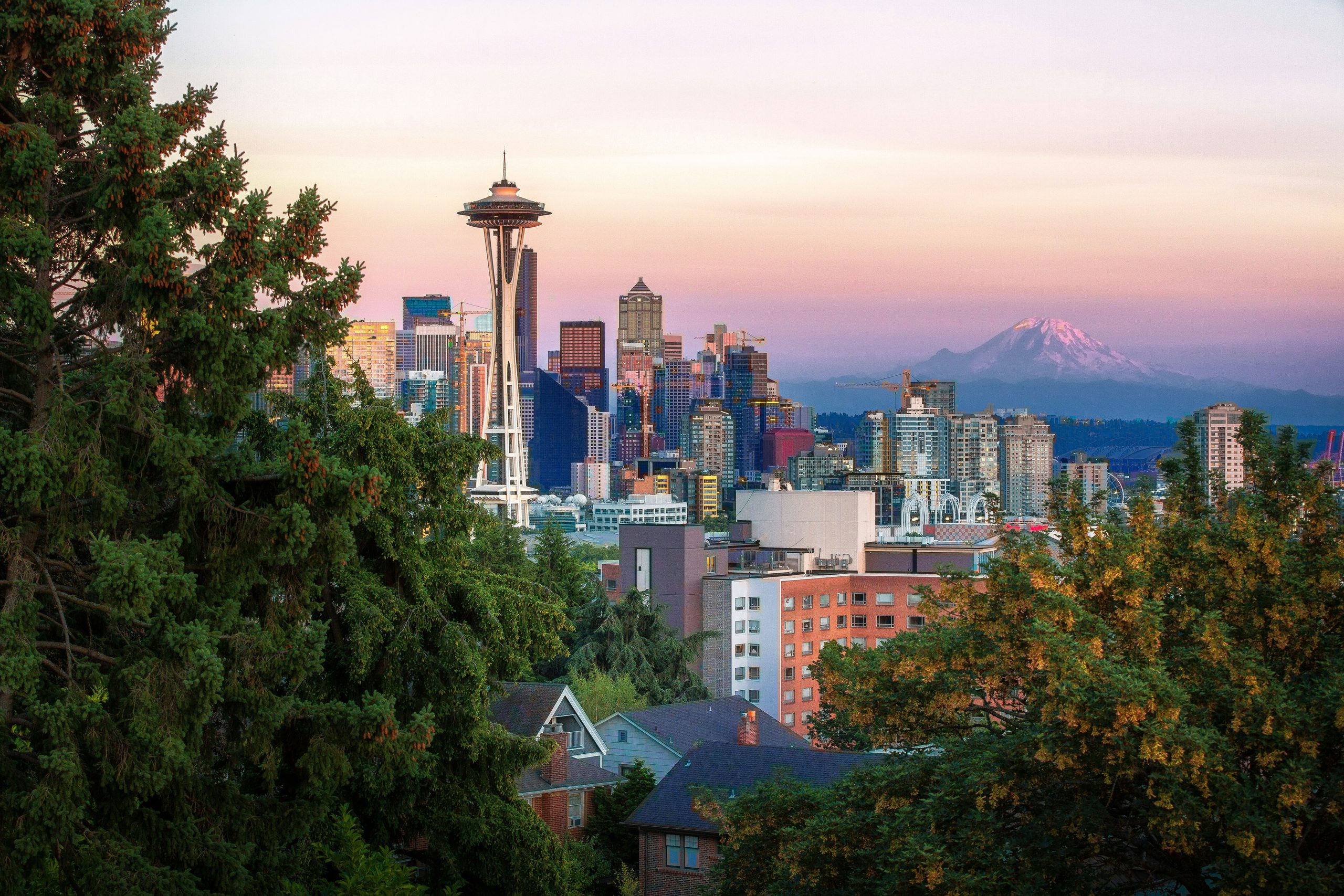 The Seattle skyline at sunset with the Space Needle, downtown skyscrapers, and Mount Rainier in the distance.
